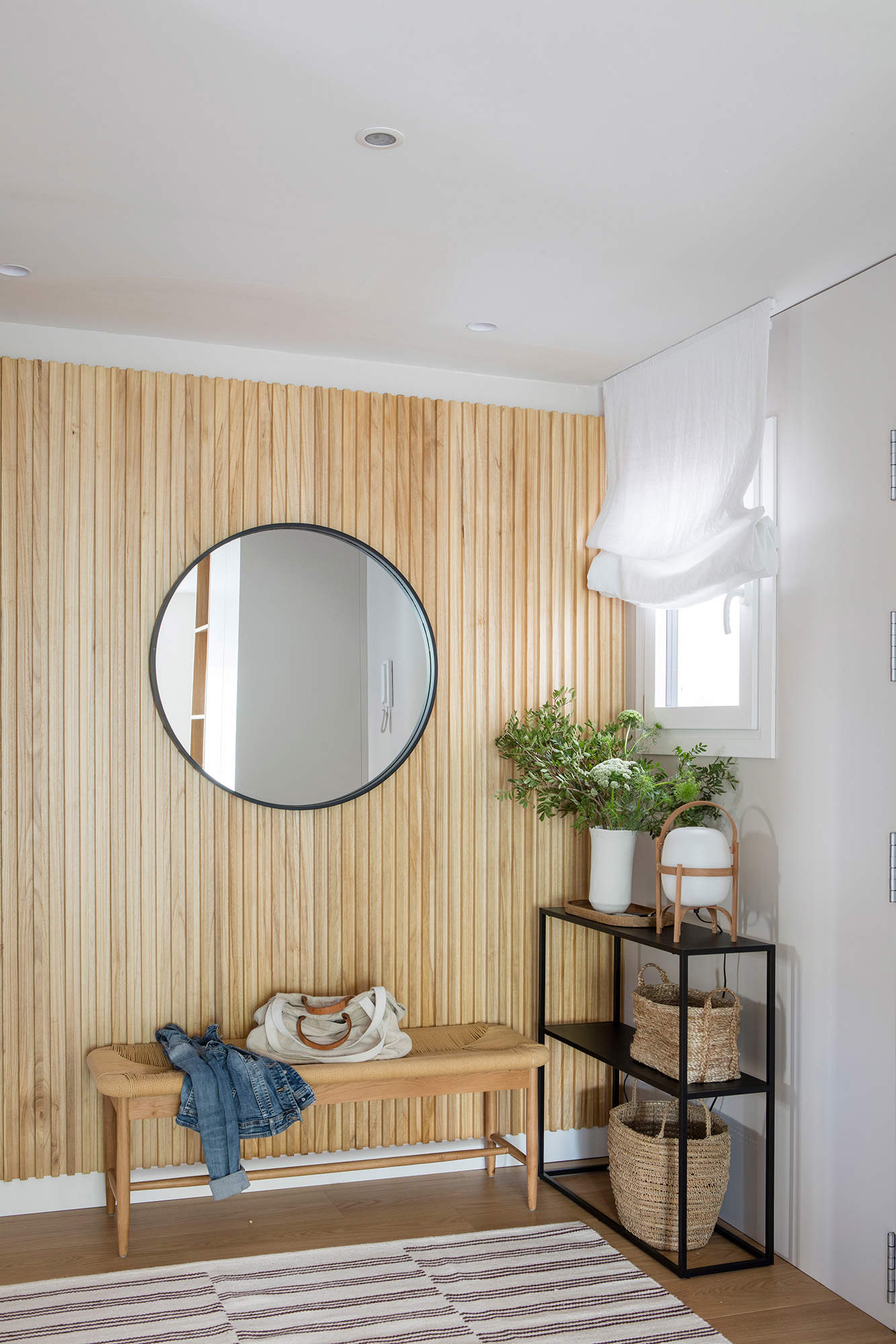 Recibidor con pared de listones de madera. Espejo redondo y banco de fibras naturales y patas de madera. Al costado, una estantería de metal negra.