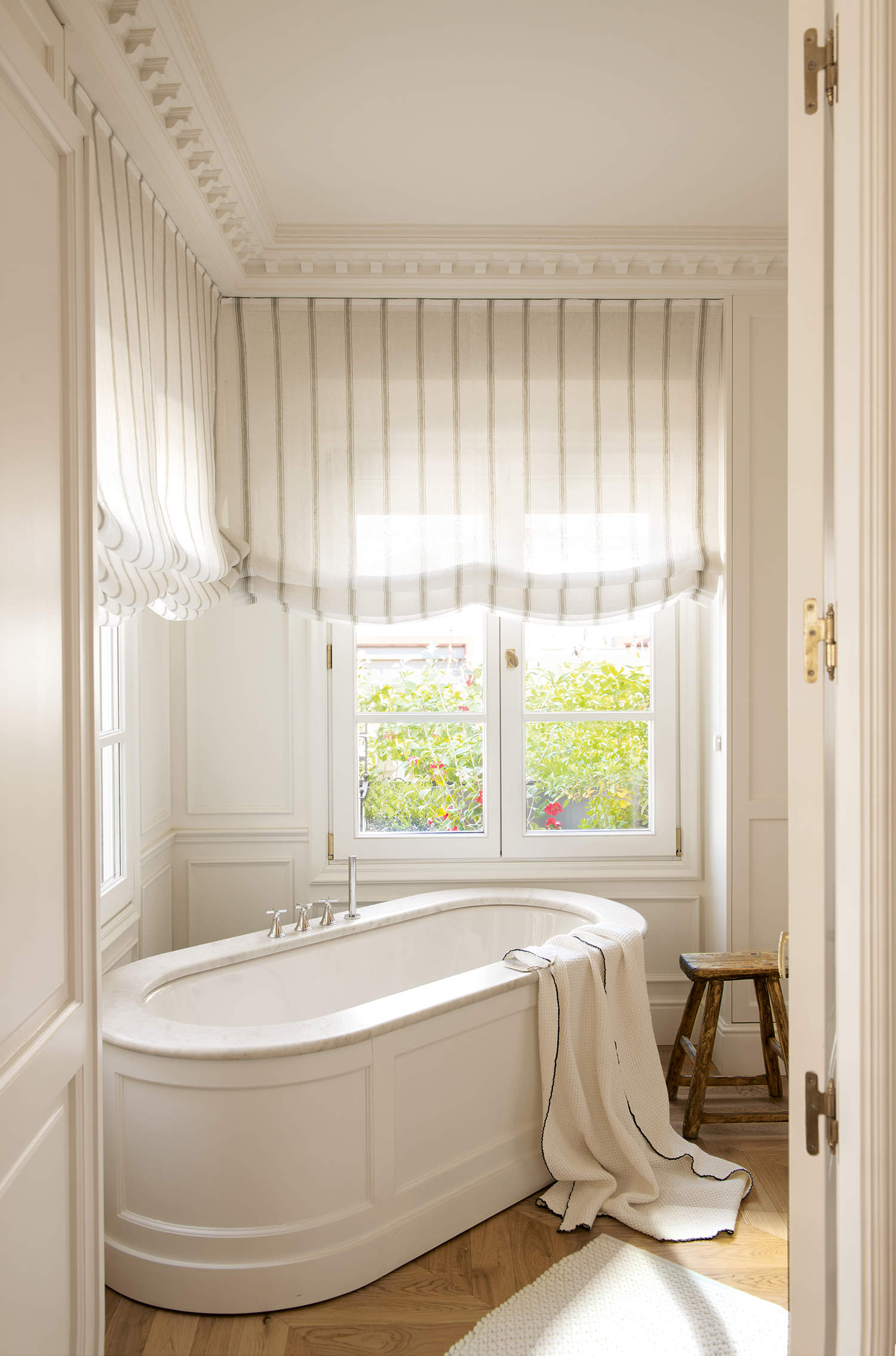 Baño elegante y clásico con bañera, suelo de madera y estores a rayas