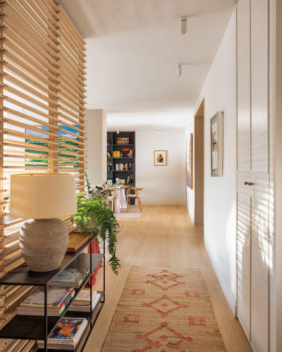 Listones de madera en horizontal en recibidor pequeño. Pasillo con alfombra de tonos beige y rojos y una estantería de metal negro con libros y lámpara gris con pantalla color crema. 