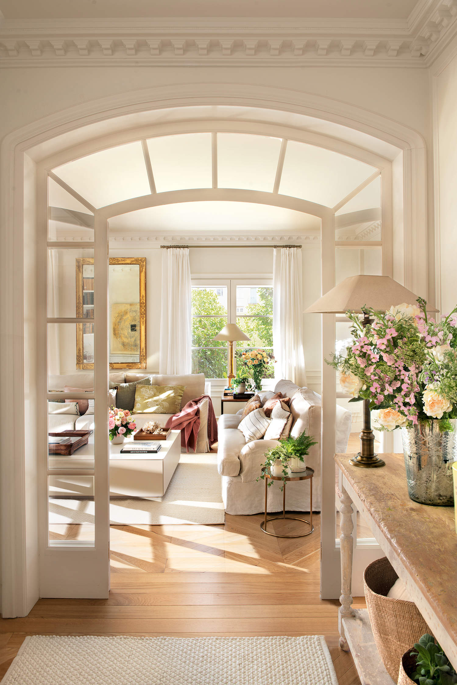 Recibidor clásico con salón al fondo, puerta con arco de cristal, sofás blancos y flores