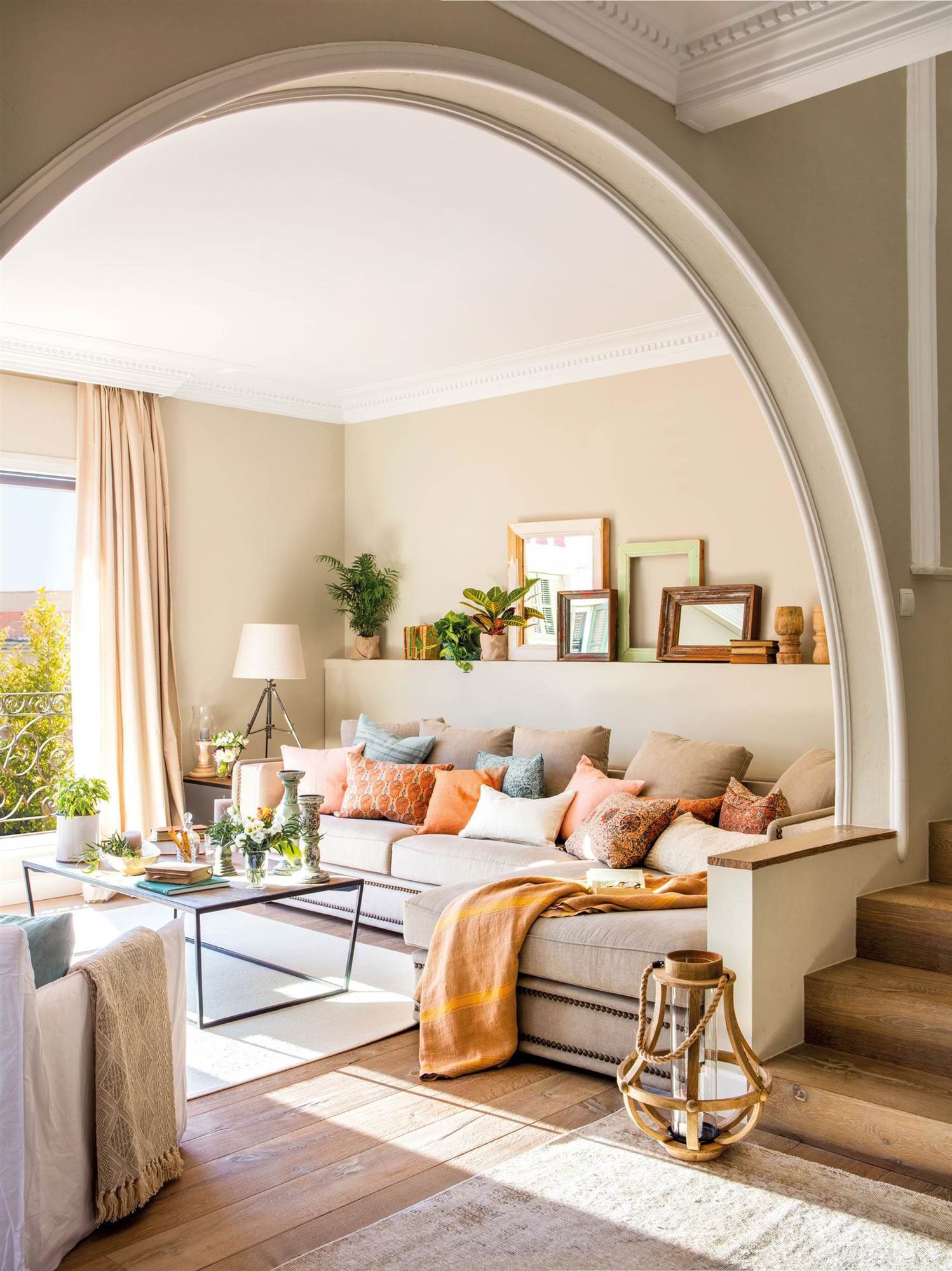 Salón con arco en la entrada, sofá beige con puff a juego, cojines, estante con cuadros y mesa de centro,.